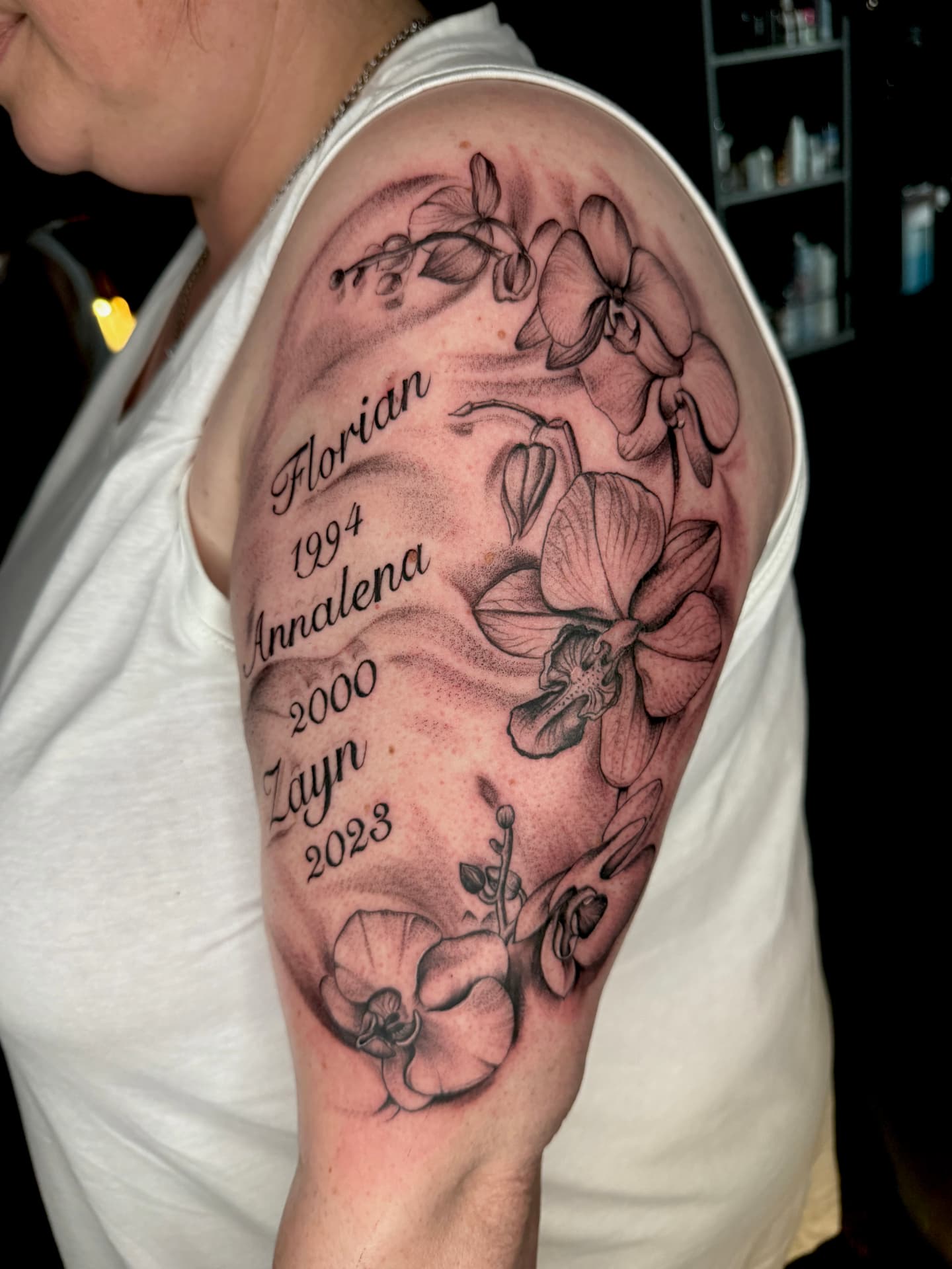 Orchideen-Tattoo mit Namen und Jahreszahlen am Oberarm in Black & Grey